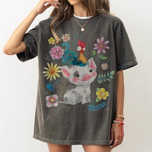 Pu&ograve; includere: T-shirt grigio scuro con un maiale dei cartoni animati e un gallo sulla testa, circondato da fiori e foglie colorate. Il maiale sorride e il gallo ha una cresta rossa. La maglietta ha un aspetto vintage.