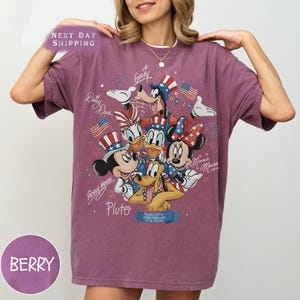 May include: Berry-coloured t-shirt featuring a graphic of Disney characters Mickey Mouse, Minnie Mouse, Donald Duck, Daisy Duck, Goofy, and Pluto, with American flags and fireworks. The shirt has the text "Mickey Mouse", "Pluto", and "USA Anniversary 1776-2024".