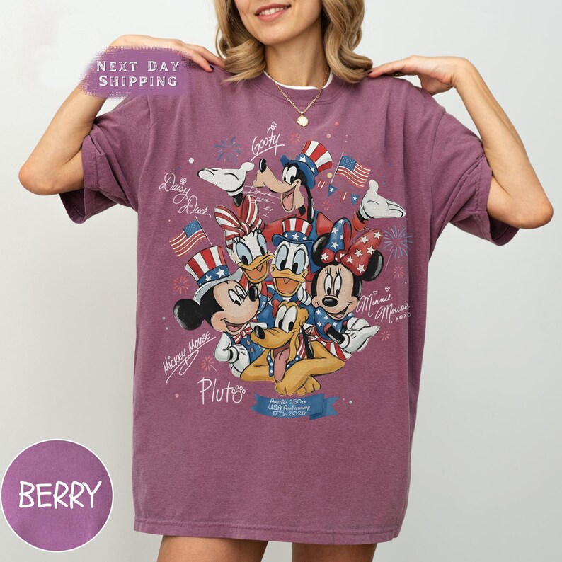May include: Berry-coloured t-shirt featuring a graphic of Disney characters Mickey Mouse, Minnie Mouse, Donald Duck, Daisy Duck, Goofy, and Pluto, with American flags and fireworks. The shirt has the text "Mickey Mouse", "Pluto", and "USA Anniversary 1776-2024".