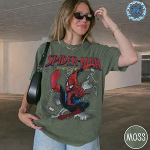 May include: An olive green t-shirt with the words "SPIDER-MAN" in red and blue, and a graphic of Spider-Man. The shirt also has the text "BRAND NEW DAY". The t-shirt is worn by a person wearing sunglasses.