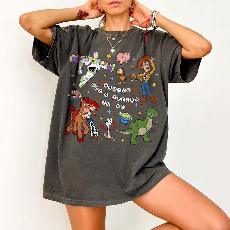 May include: Dark grey t-shirt with a colourful graphic of characters from the animated film "Toy Story." The design includes Buzz Lightyear, Woody, and other characters, with the phrase "You've Got a Friend in Me."