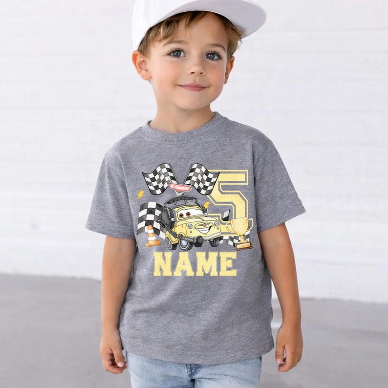 Pode incluir: Uma camiseta cinza com um carro de desenho animado, bandeiras quadriculadas, o n&uacute;mero 5 e a palavra "NAME" em amarelo. A camiseta &eacute; usada por uma crian&ccedil;a pequena que usa um bon&eacute; de beisebol branco.