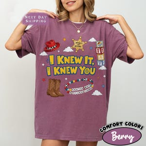 Pode incluir: Uma t-shirt cor de baga com uma impress&atilde;o gr&aacute;fica de um chap&eacute;u de cowboy vermelho, uma estrela de xerife dourada, blocos de letras, botas de cowboy castanhas e uma pulseira de contas. O texto diz I KNEW IT, I KNEW YOU, INFINITY AND BEYOND e YOU'VE GOT A FRIEND IN ME.