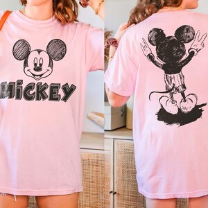 May include: Pale pink t-shirt with a black and white Mickey Mouse design on the front and back. The front graphic shows Mickey's face and the word "Mickey". The back graphic shows Mickey with his arms raised.