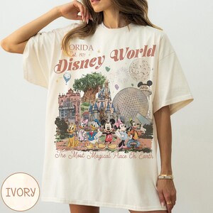 K&ouml;nnte beinhalten: Elfenbeinfarbenes T-Shirt mit einem Disney World-Motiv. Das Design enth&auml;lt den Text "Florida Est. 1971" und "Disney World" in einer Vintage-Schriftart sowie Illustrationen von Disney-Figuren und Park-Wahrzeichen. Der Satz "The Most Magical Place On Earth" ist ebenfalls vorhanden.