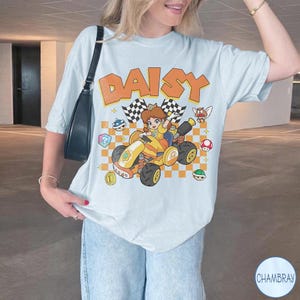 May include: A light blue t-shirt featuring a graphic print of the character Daisy driving a yellow race car. The design includes the text DAISY in orange letters, a checkered background, and various game icons like shells, a mushroom, and a coin.