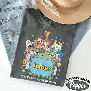 May include: A charcoal gray t-shirt featuring a graphic print of Toy Story 5 characters surrounding a large green frog-shaped frame with the name James printed in yellow. The text You've Got A Friend In Me is printed below the frog frame.