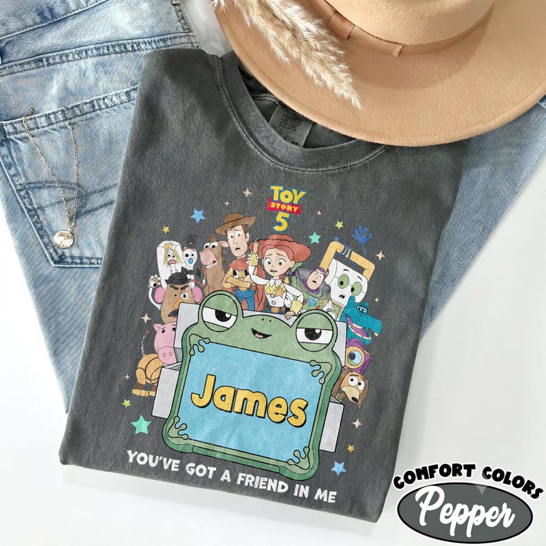 May include: A charcoal gray t-shirt featuring a graphic print of Toy Story 5 characters surrounding a large green frog-shaped frame with the name James printed in yellow. The text You've Got A Friend In Me is printed below the frog frame.