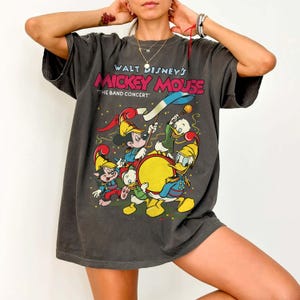 May include: Dark gray t-shirt featuring a colorful graphic of Mickey Mouse and friends playing musical instruments. The text on the shirt reads "Walt Disney's Mickey Mouse" and "The Band Concert". The shirt has short sleeves and a relaxed fit.