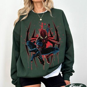Puede incluir: Sudadera verde bosque con un dise&ntilde;o de Spider-Man. El dise&ntilde;o incluye una telara&ntilde;a roja y negra y salpicaduras rojas. La sudadera tiene cuello redondo y mangas largas. El texto "Forest Green" est&aacute; en la parte inferior.