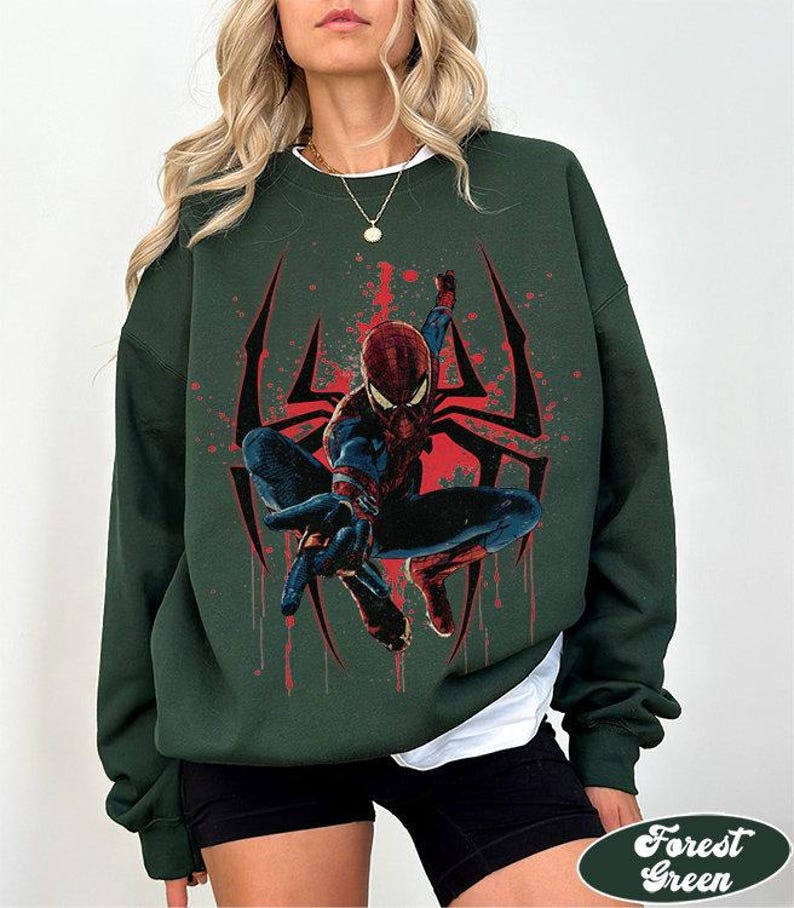 Puede incluir: Sudadera verde bosque con un dise&ntilde;o de Spider-Man. El dise&ntilde;o incluye una telara&ntilde;a roja y negra y salpicaduras rojas. La sudadera tiene cuello redondo y mangas largas. El texto "Forest Green" est&aacute; en la parte inferior.