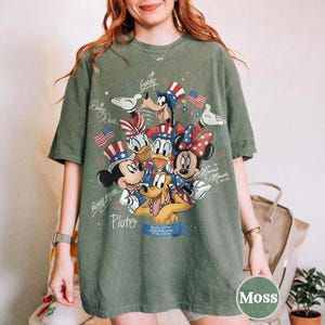 May include: A moss green t-shirt with a graphic of Disney characters Mickey Mouse, Minnie Mouse, Donald Duck, Daisy Duck, Goofy, and Pluto, in patriotic outfits. The design includes American flags and fireworks.