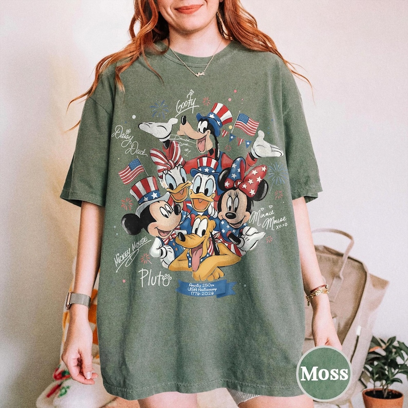 May include: A moss green t-shirt with a graphic of Disney characters Mickey Mouse, Minnie Mouse, Donald Duck, Daisy Duck, Goofy, and Pluto, in patriotic outfits. The design includes American flags and fireworks.