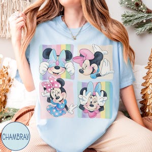 May include: Light blue t-shirt featuring a four-panel graphic of Minnie Mouse in various poses, each framed with pastel rainbow stripes. Minnie is wearing different colored bows and dresses. The shirt has the word "CHAMBRAY" printed on it.