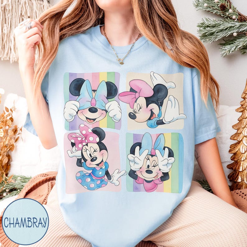 May include: Light blue t-shirt featuring a four-panel graphic of Minnie Mouse in various poses, each framed with pastel rainbow stripes. Minnie is wearing different colored bows and dresses. The shirt has the word "CHAMBRAY" printed on it.