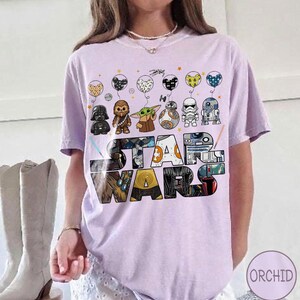 May include: A lilac t-shirt with the words "STAR WARS" in a colourful, blocky design. Above the text are cartoon character balloons. The shirt has short sleeves and a crew neck.