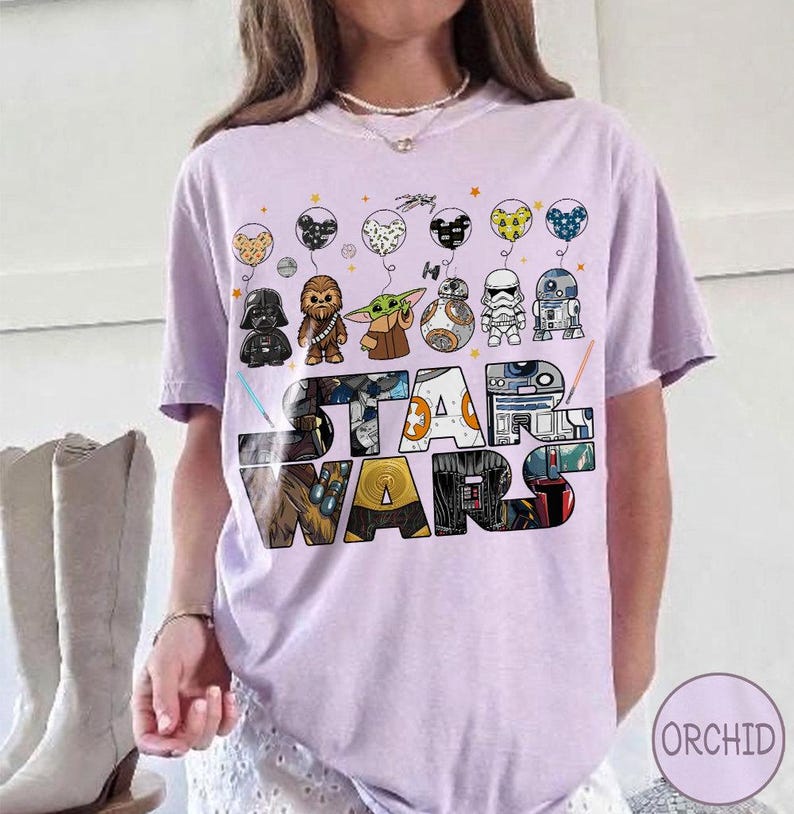 May include: A lilac t-shirt with the words "STAR WARS" in a colourful, blocky design. Above the text are cartoon character balloons. The shirt has short sleeves and a crew neck.