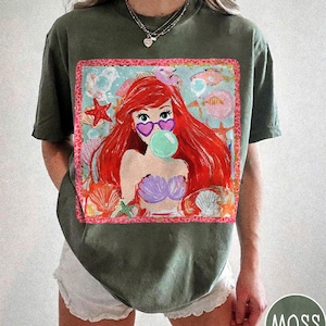 Puede incluir: Camiseta verde musgo con un gr&aacute;fico colorido de una sirena con pelo rojo, gafas de sol en forma de coraz&oacute;n y una burbuja. El dise&ntilde;o incluye conchas marinas, estrellas de mar y peces. La camiseta es de corte holgado.