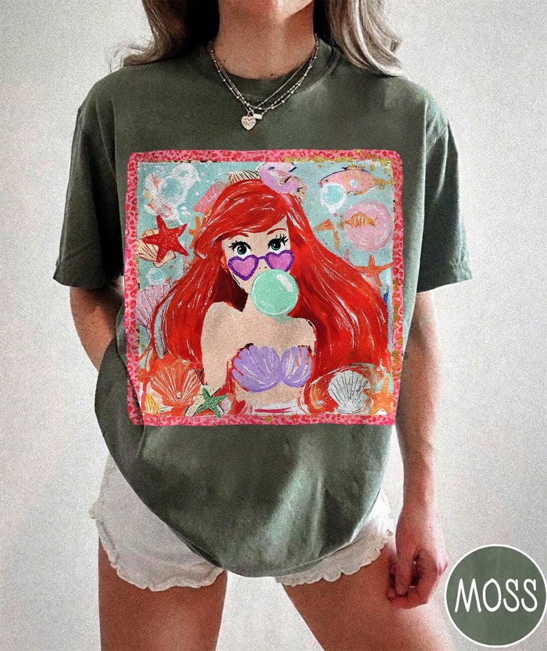 Puede incluir: Camiseta verde musgo con un gr&aacute;fico colorido de una sirena con pelo rojo, gafas de sol en forma de coraz&oacute;n y una burbuja. El dise&ntilde;o incluye conchas marinas, estrellas de mar y peces. La camiseta es de corte holgado.