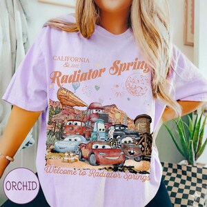 May include: A light purple t-shirt featuring a graphic of characters from the movie Cars in Radiator Springs. The shirt has the text "CALIFORNIA Est. 2012" and "Radiator Springs" and "Welcome to Radiator Springs".