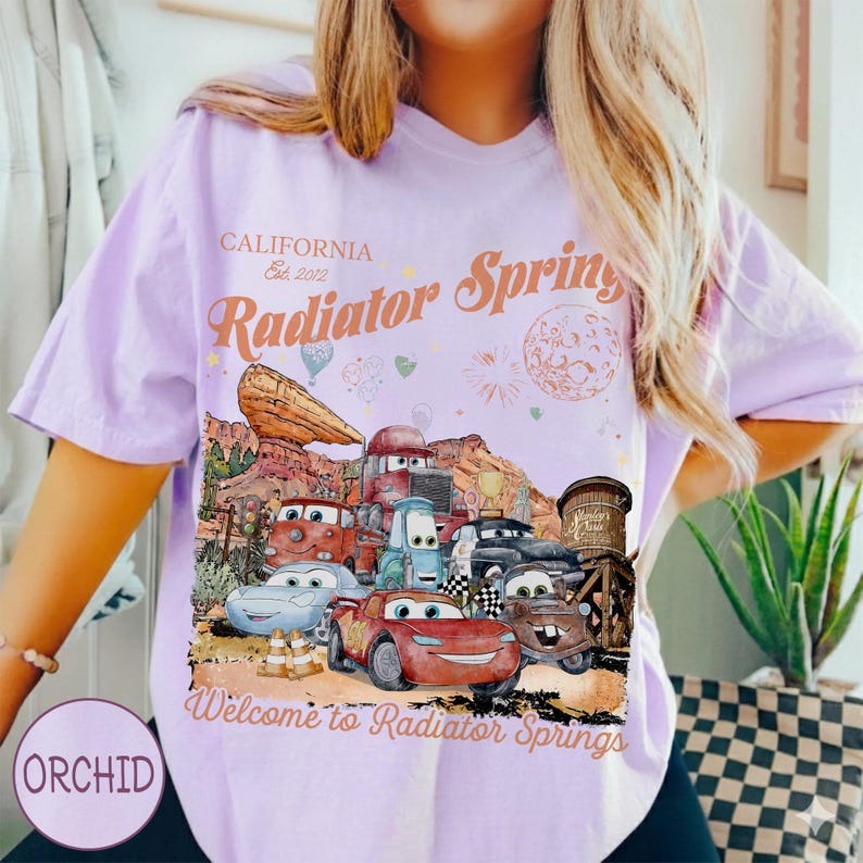 May include: A light purple t-shirt featuring a graphic of characters from the movie Cars in Radiator Springs. The shirt has the text "CALIFORNIA Est. 2012" and "Radiator Springs" and "Welcome to Radiator Springs".