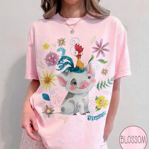 Pu&ograve; includere: T-shirt rosa chiaro con un maiale dei cartoni animati con un gallo in testa, circondato da fiori e foglie colorate. La parola "Blossom" &egrave; in un cerchio in basso a destra sulla maglietta.