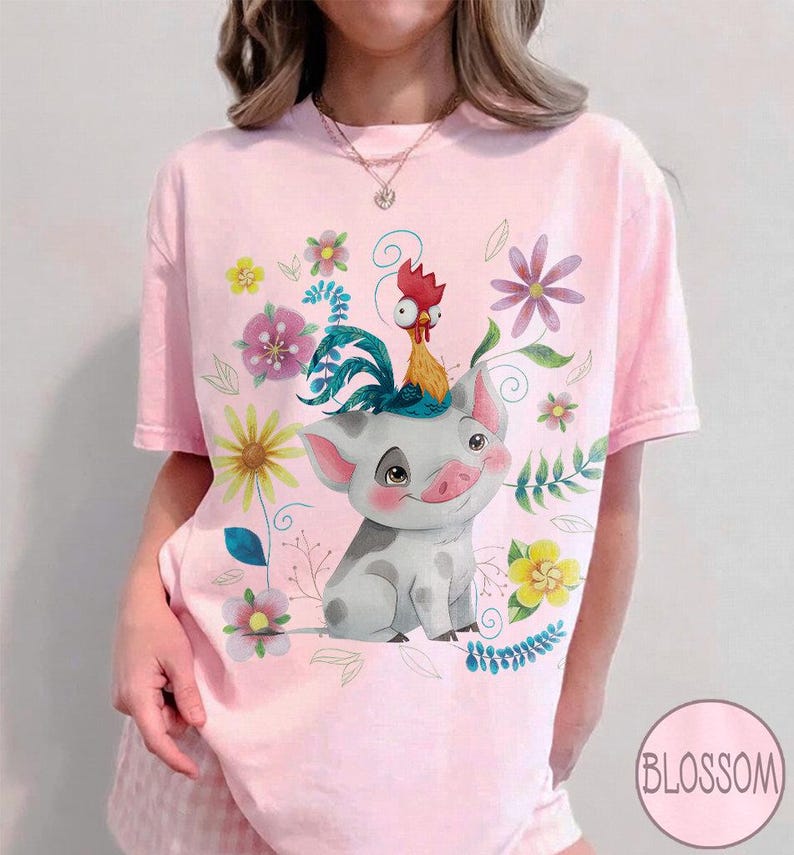 Pu&ograve; includere: T-shirt rosa chiaro con un maiale dei cartoni animati con un gallo in testa, circondato da fiori e foglie colorate. La parola "Blossom" &egrave; in un cerchio in basso a destra sulla maglietta.