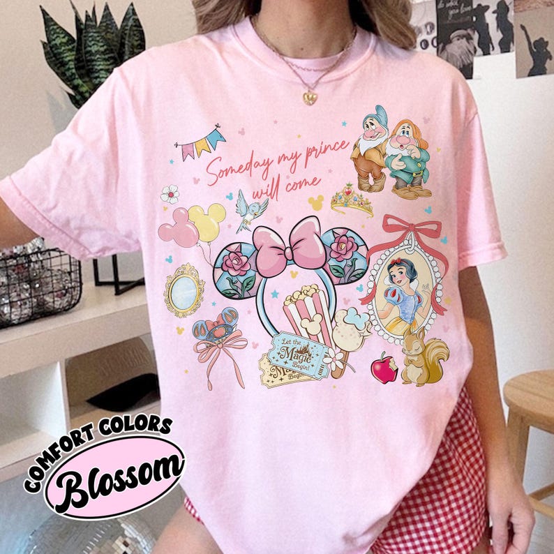 May include: Pink t-shirt with a graphic featuring Snow White, the Seven Dwarfs, and Mickey Mouse ears. The design includes the text "Someday my prince will come" and "Let the Magic Begin". The shirt is from the Comfort Colors Blossom collection.