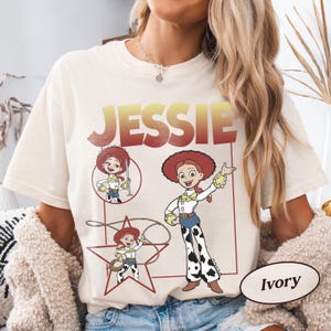 May include: Ivory-colored t-shirt featuring a graphic of the character Jessie from Toy Story. The design includes the name "JESSIE" in a gradient red and yellow font, along with multiple illustrations of the character in various poses. The shirt is a relaxed fit.