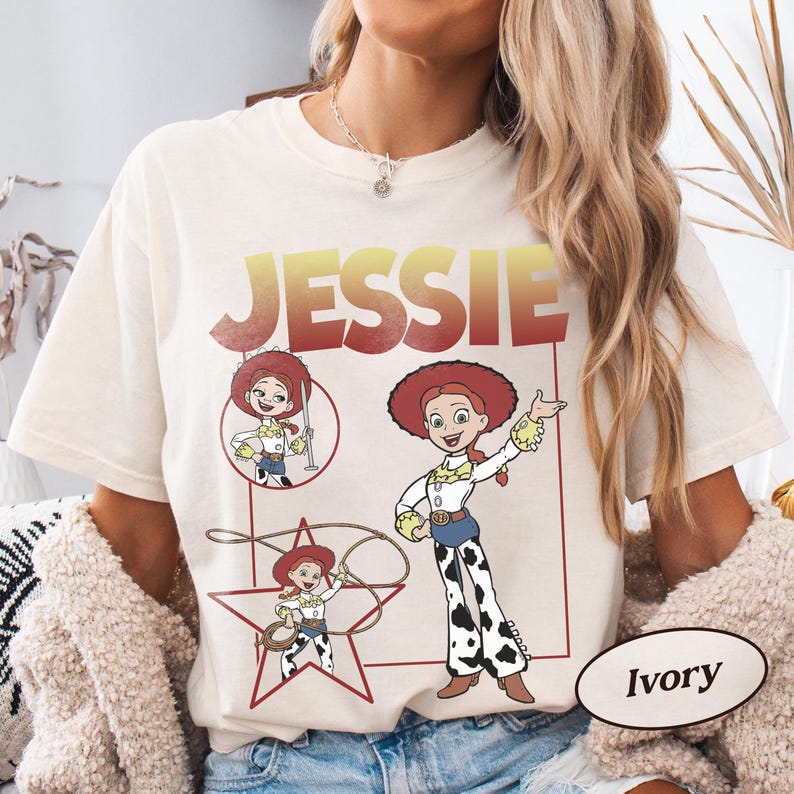 May include: Ivory-colored t-shirt featuring a graphic of the character Jessie from Toy Story. The design includes the name "JESSIE" in a gradient red and yellow font, along with multiple illustrations of the character in various poses. The shirt is a relaxed fit.