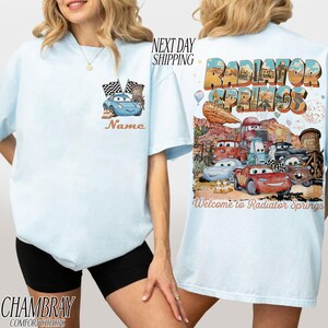 Puede incluir: Camiseta azul claro con un coche de dibujos animados y banderas en la parte delantera, con la palabra "Name". La parte trasera presenta "Radiator Springs" con personajes de coches y la frase "Welcome to Radiator Springs".