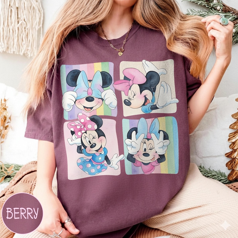 May include: Berry-colored t-shirt featuring four squares with Minnie Mouse illustrations. Each square showcases a different pose of Minnie Mouse with colorful backgrounds. The shirt has short sleeves and a round neckline.