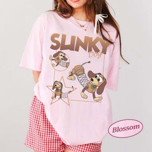 May include: Light pink t-shirt featuring the text "SLINKY Dog" in brown, with cartoon images of Slinky Dog. The shirt has short sleeves and a relaxed fit. The bottom of the shirt is paired with red and white checkered shorts.
