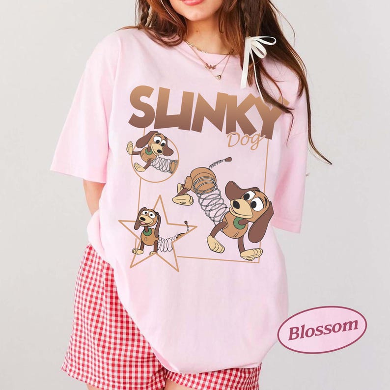 May include: Light pink t-shirt featuring the text "SLINKY Dog" in brown, with cartoon images of Slinky Dog. The shirt has short sleeves and a relaxed fit. The bottom of the shirt is paired with red and white checkered shorts.