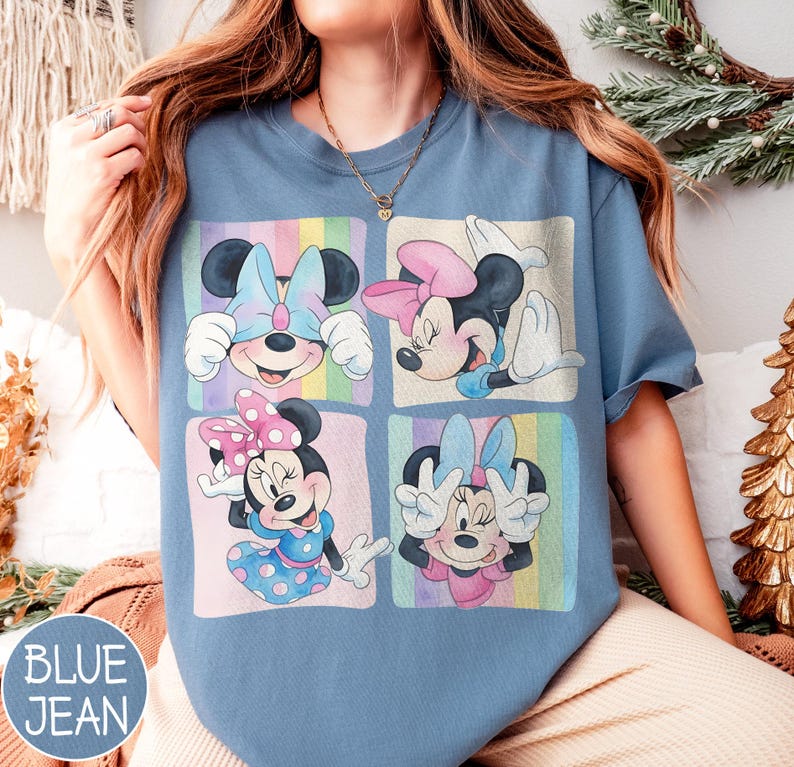 May include: Blue jean colored t-shirt featuring four panels with Minnie Mouse illustrations. Each panel showcases Minnie in different poses, wearing bows and dresses in pastel colors. The shirt has a relaxed fit.