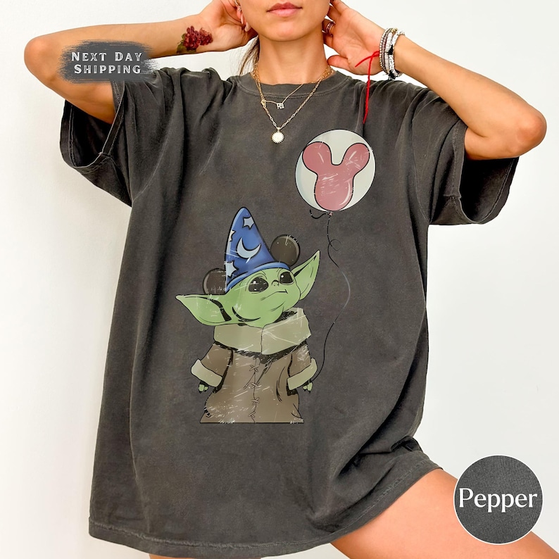 Puede incluir: Camiseta gris oscuro con un personaje verde que lleva un sombrero de mago azul con orejas de Mickey Mouse, sosteniendo un globo de Mickey Mouse. La camiseta tiene el texto "Next Day Shipping" y el color es "Pepper".