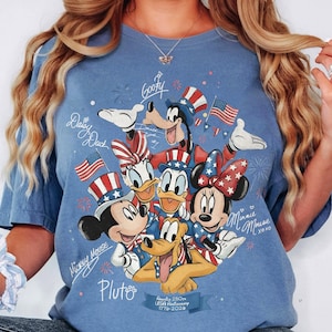 May include: Blue t-shirt with a graphic of Disney characters Mickey Mouse, Minnie Mouse, Donald Duck, Daisy Duck, Goofy, and Pluto, wearing patriotic hats and holding flags. Text includes "Mickey Mouse", "Minnie Mouse", "Daisy Duck", "Goofy", and "Pluto".