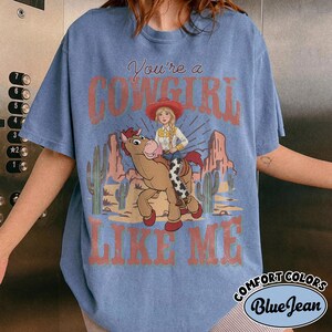 May include: A blue short-sleeve t-shirt featuring a graphic print of a person with blonde hair wearing a red cowboy hat and riding a brown horse. The text reads You're a COWGIRL LIKE ME. The background shows a desert landscape with cacti.