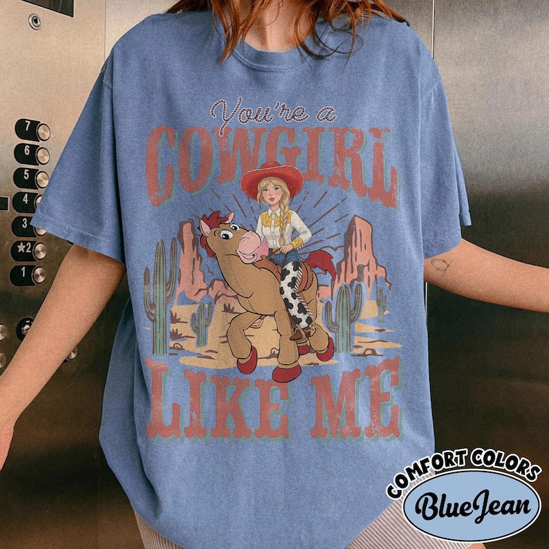 May include: A blue short-sleeve t-shirt featuring a graphic print of a person with blonde hair wearing a red cowboy hat and riding a brown horse. The text reads You're a COWGIRL LIKE ME. The background shows a desert landscape with cacti.