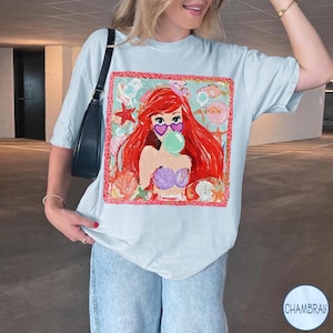 Puede incluir: Camiseta azul claro con un gr&aacute;fico colorido de una sirena de dibujos animados con cabello rojo, gafas de sol en forma de coraz&oacute;n y una burbuja de chicle. El gr&aacute;fico est&aacute; enmarcado con un borde rojo y elementos tem&aacute;ticos marinos.
