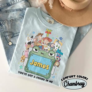 May include: A light blue t-shirt featuring a Toy Story 5 graphic with various characters, a green frog-shaped frame containing the name James, and the text You've got a friend in me. The shirt has a Comfort Colors label at the inner neckline.