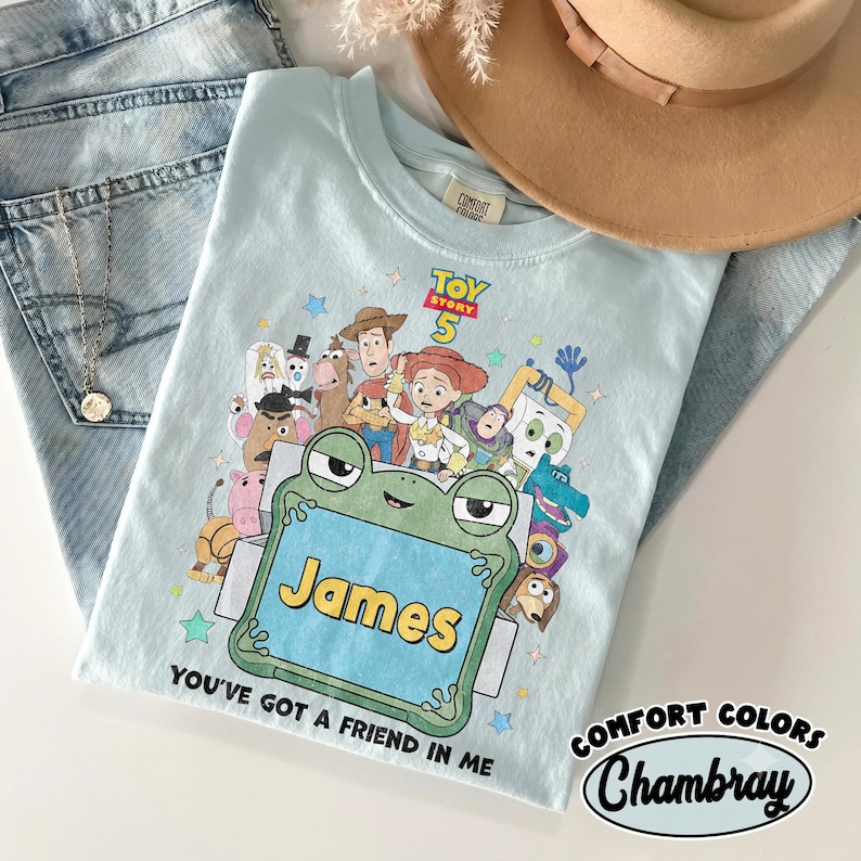 May include: A light blue t-shirt featuring a Toy Story 5 graphic with various characters, a green frog-shaped frame containing the name James, and the text You've got a friend in me. The shirt has a Comfort Colors label at the inner neckline.