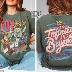 K&ouml;nnte beinhalten: Olivgr&uuml;nes T-Shirt mit den Worten "Toy Story" in Rosa und Wei&szlig;, mit Grafiken von Woody und Buzz Lightyear. Auf der R&uuml;ckseite des Shirts steht "To Infinity and Beyond" mit Buzz & Woody. Das Shirt ist im Comfort-Colors-Stil.