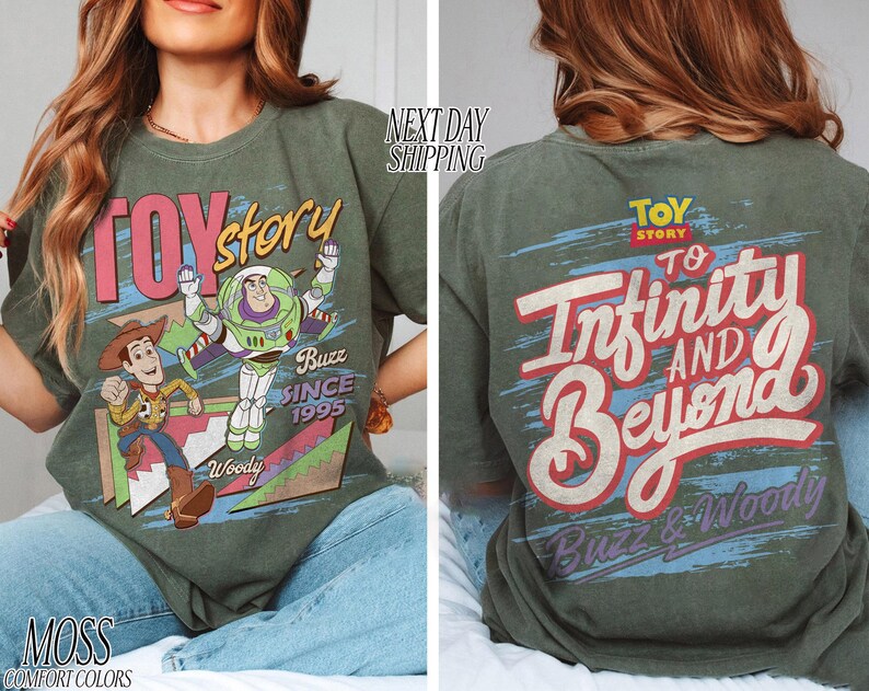 K&ouml;nnte beinhalten: Olivgr&uuml;nes T-Shirt mit den Worten "Toy Story" in Rosa und Wei&szlig;, mit Grafiken von Woody und Buzz Lightyear. Auf der R&uuml;ckseite des Shirts steht "To Infinity and Beyond" mit Buzz & Woody. Das Shirt ist im Comfort-Colors-Stil.