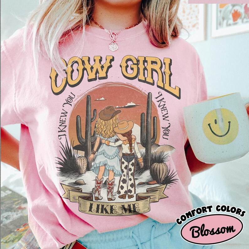 May include: A light pink t-shirt featuring a graphic of two people in cowboy hats standing in a desert with cacti. The text reads COW GIRL at the top and I KNEW YOU I KNEW YOU LIKE ME around the illustration. The design includes a sunset background.