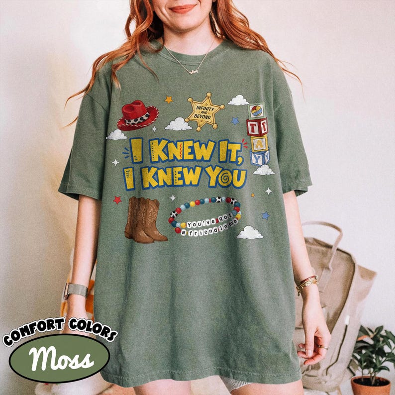 Pode incluir: Uma t-shirt verde musgo com uma estampa gr&aacute;fica que inclui um chap&eacute;u de cowboy vermelho, uma estrela de xerife dourada, blocos de alfabeto, botas de cowboy castanhas e uma pulseira de contas. O texto diz I KNEW IT, I KNEW YOU e YOU'VE GOT A FRIEND IN ME.