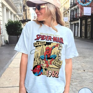 May include: Light blue t-shirt featuring a vintage Spider-Man graphic. The design includes the words "BRAND NEW DAY SPIDER-MAN NEW YORK" and "1962". The graphic depicts Spider-Man in action with a cityscape background. The shirt is a casual wear item.
