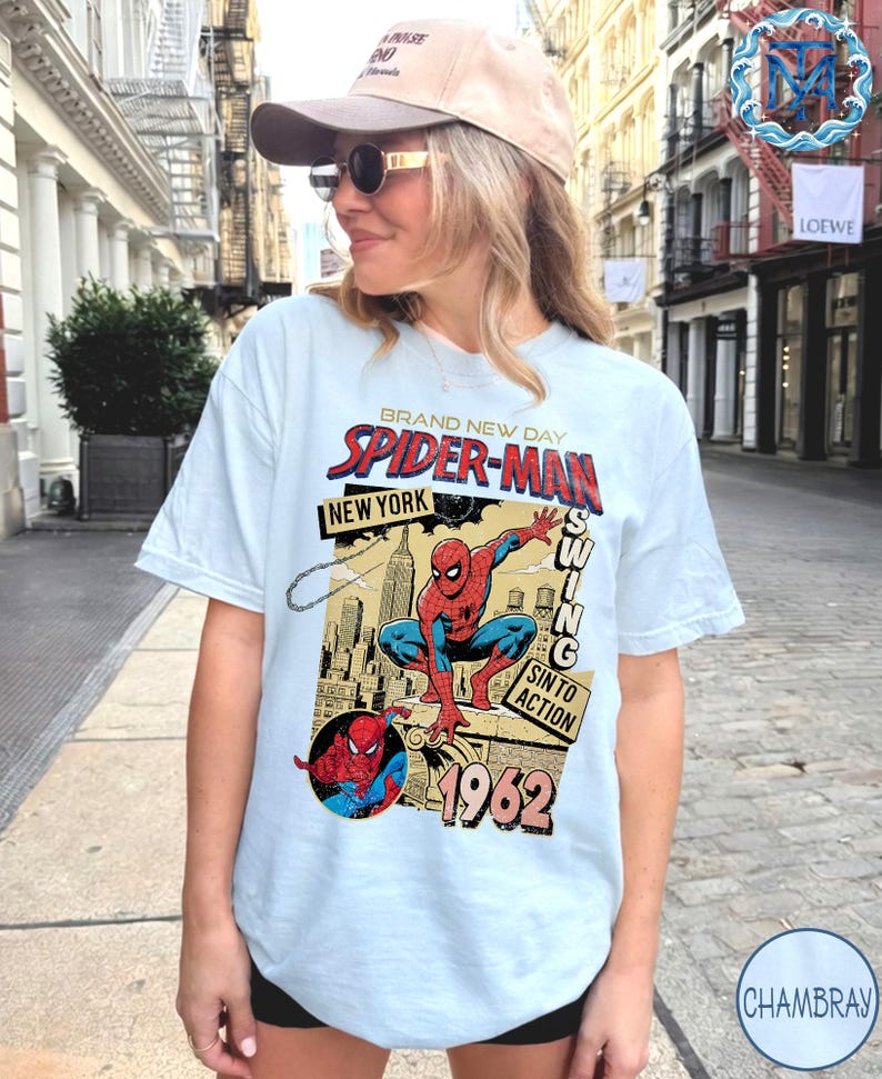 May include: Light blue t-shirt featuring a vintage Spider-Man graphic. The design includes the words "BRAND NEW DAY SPIDER-MAN NEW YORK" and "1962". The graphic depicts Spider-Man in action with a cityscape background. The shirt is a casual wear item.