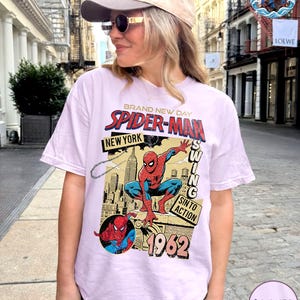 May include: A light purple t-shirt featuring a vintage Spider-Man graphic. The design includes the words "BRAND NEW DAY," "SPIDER-MAN," "NEW YORK," and "1962." The graphic depicts Spider-Man in action with a cityscape background.