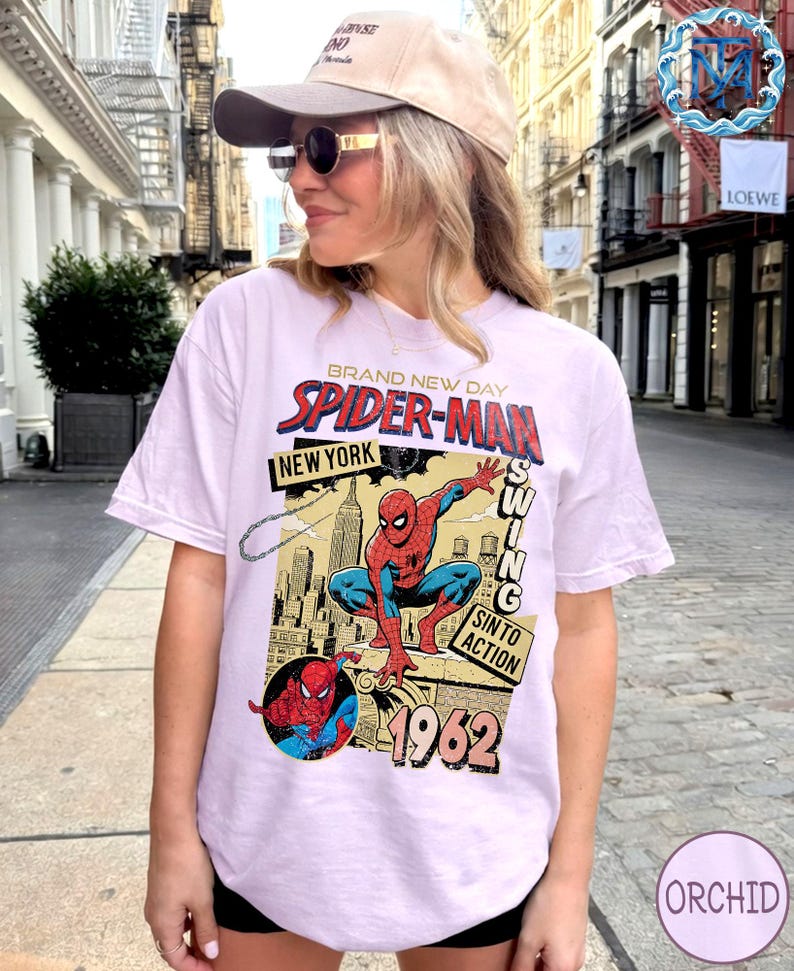 May include: A light purple t-shirt featuring a vintage Spider-Man graphic. The design includes the words "BRAND NEW DAY," "SPIDER-MAN," "NEW YORK," and "1962." The graphic depicts Spider-Man in action with a cityscape background.